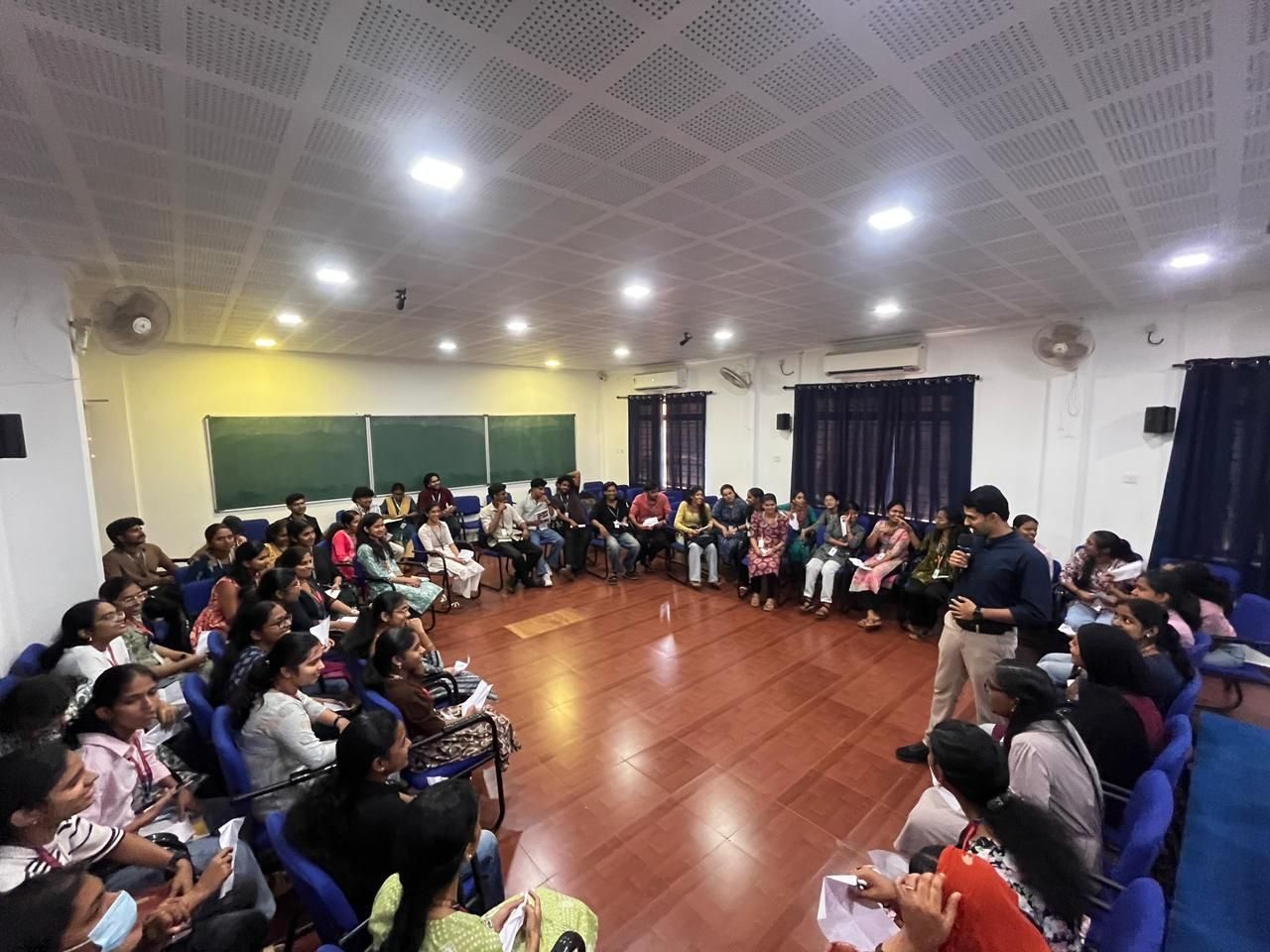 A trainer speaks to students seated in a circle around the hall at St. Joseph's College, Devagiri